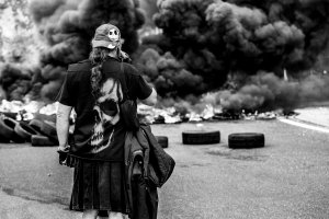 Un homme avec une casquette et un t-shirt "tête de mort" se tien debout devant un gros nuage de fumée d'un feux de pneus sur le port de Brest